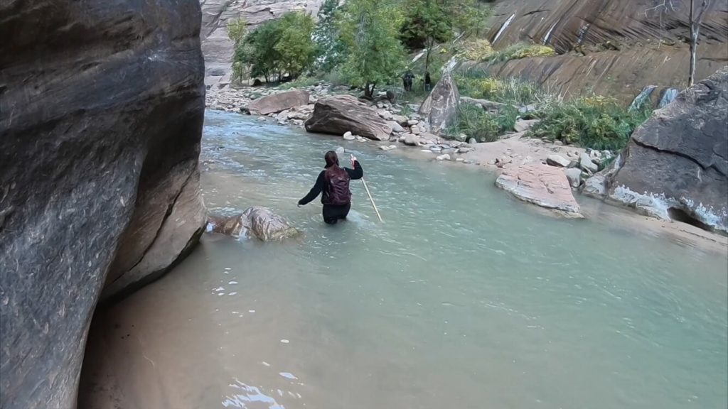 woman walking through river, water is thigh deep, hiking stick in right hand, canyon walls on both sides