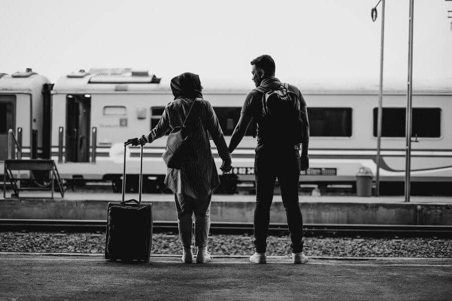 black & white photo of a couple holding hands facing train tracks. woman has a suitcase to her left